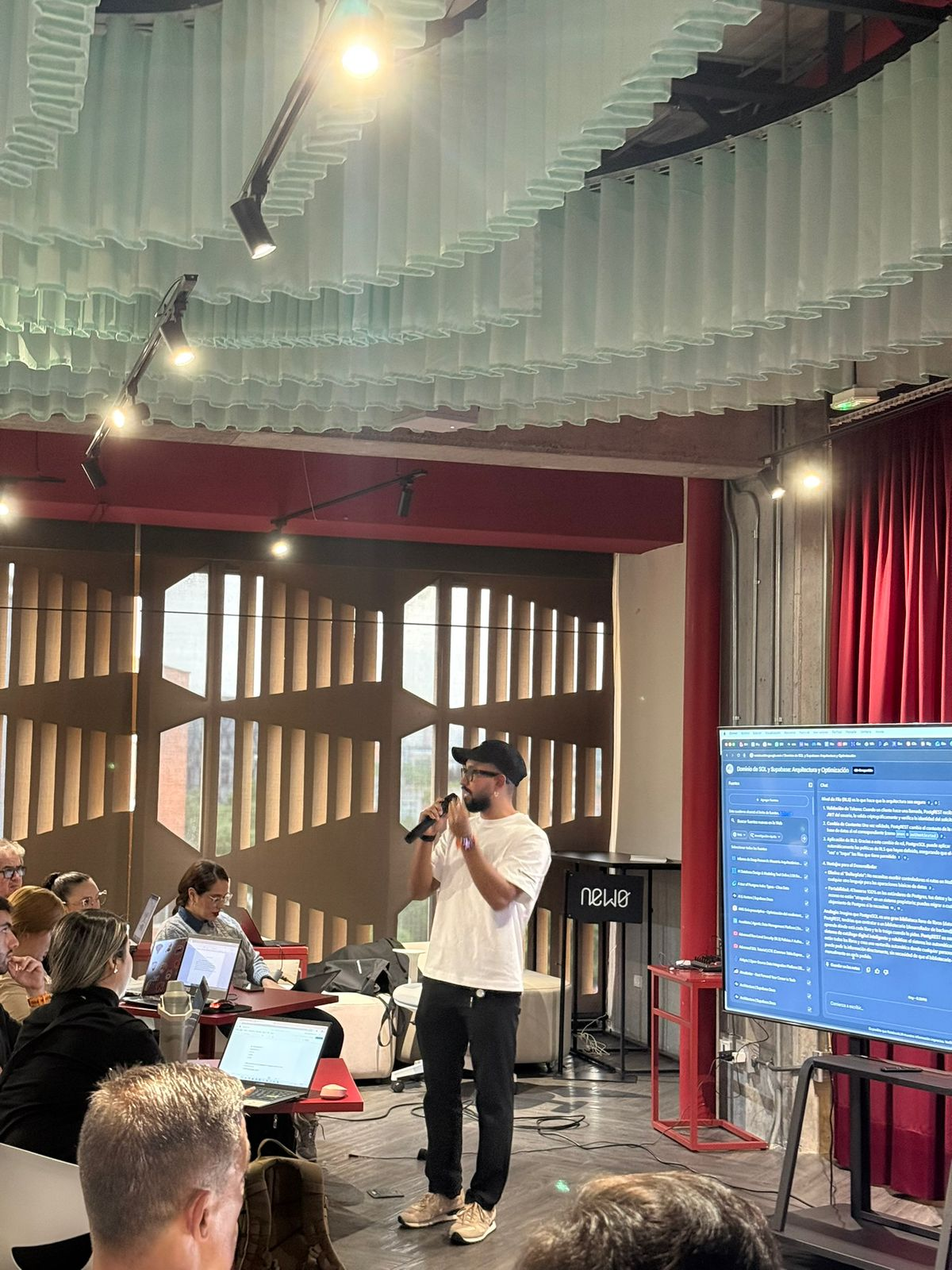 Juan Camilo Martínez, de pie con micrófono, guía una sesión sobre bases de datos mientras las personas asistentes siguen la explicación desde sus portátiles y una pantalla muestra el material de la clase.
